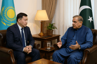 Two diplomats sit across from each other in a formal meeting room. The Kazakh representative, wearing a dark suit and tie, faces the Pakistani official, dressed in a blue traditional shalwar kameez with a waistcoat. The flags of Kazakhstan and Pakistan stand behind them, and a small table with a lamp, a plant, and a glass of water is placed between them, creating a professional and diplomatic setting.