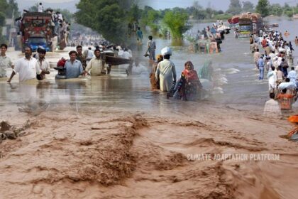 People wading through floodwaters in Pakistan as heavy monsoon rains cause severe flooding, destroying homes, displacing families, and blocking roads.