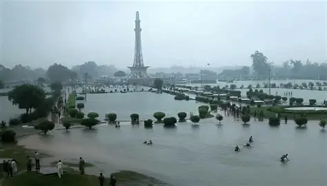 Flooded streets and heavy rain in Punjab, Pakistan during the 10th spell of monsoon rains, with people navigating through rainwater and dark stormy clouds overhead.