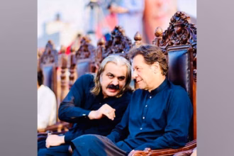 Imran Khan and Ali Amin Gandapur in traditional black attire sitting on ornate chairs, engaged in a serious conversation during a public event.