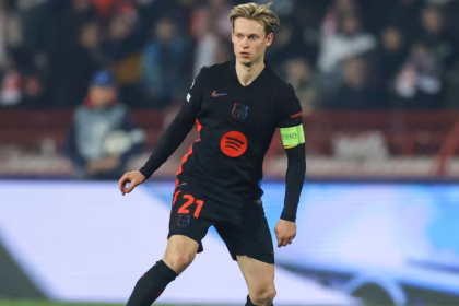 "Frenkie de Jong wearing Barcelona’s black away kit with red details and the captain’s armband, controlling the ball during a UEFA Champions League match. The stadium background is blurred with fans in the stands."