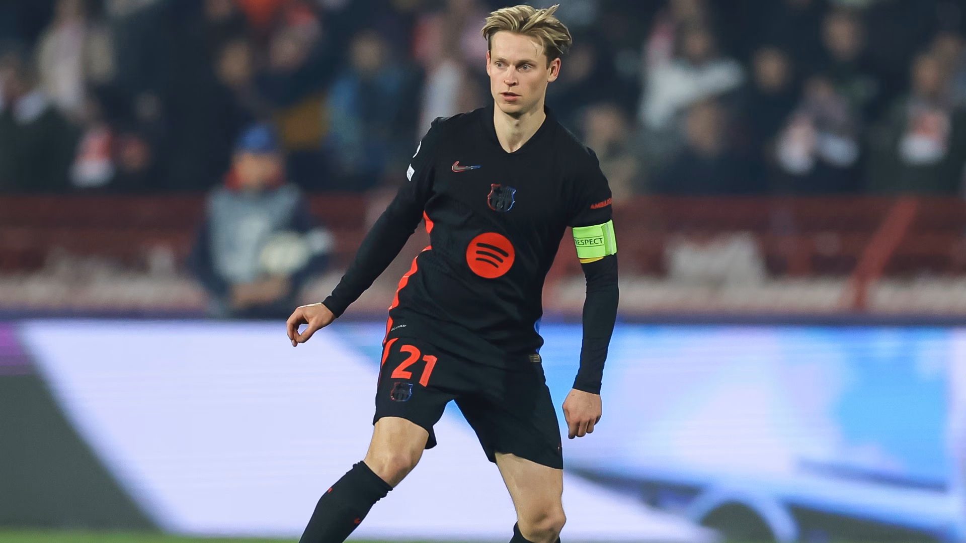 "Frenkie de Jong wearing Barcelona’s black away kit with red details and the captain’s armband, controlling the ball during a UEFA Champions League match. The stadium background is blurred with fans in the stands."